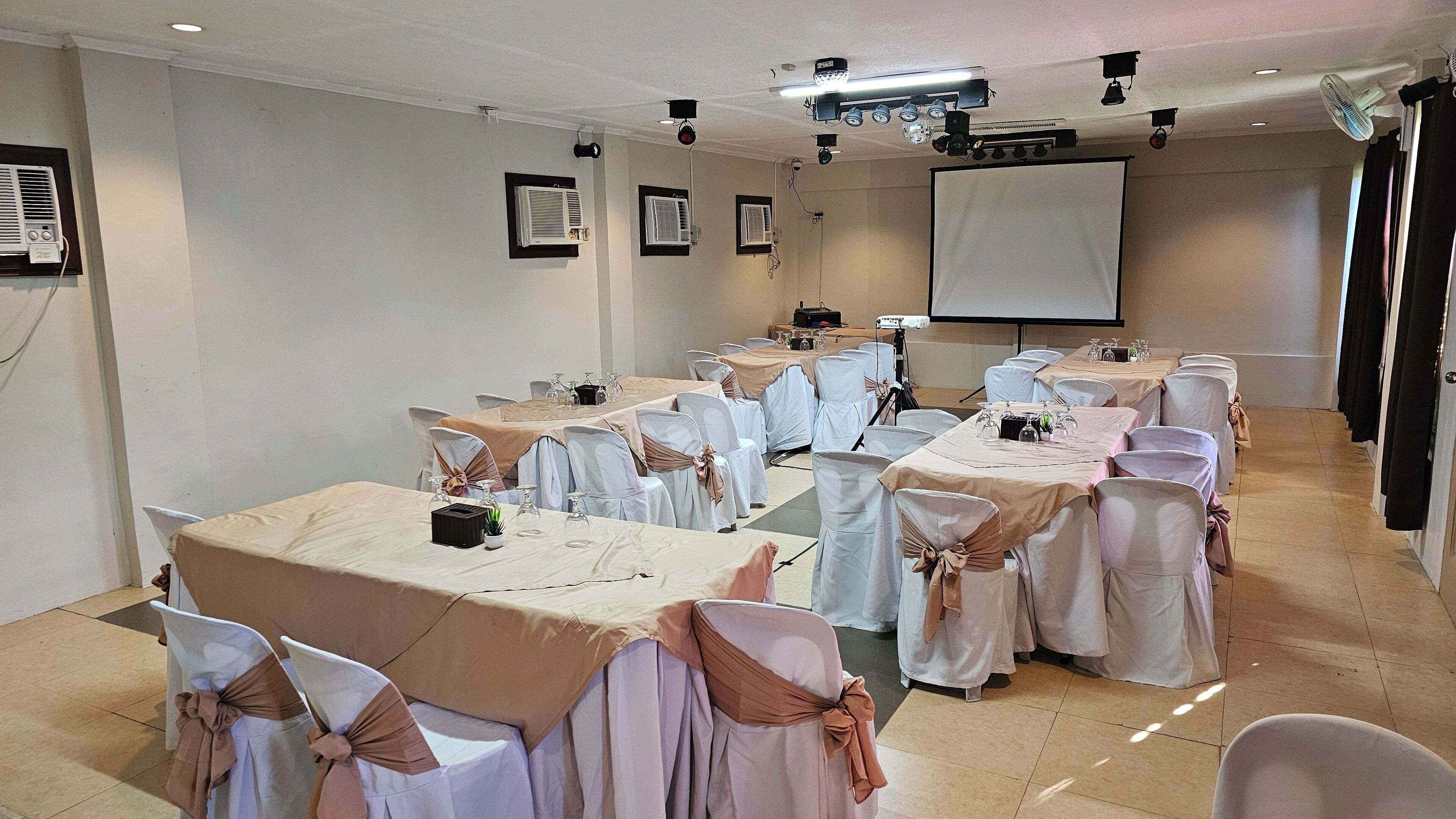 Un salón de conferencias con mesas dispuestas para una reunión, equipado con proyectores y una pantalla.