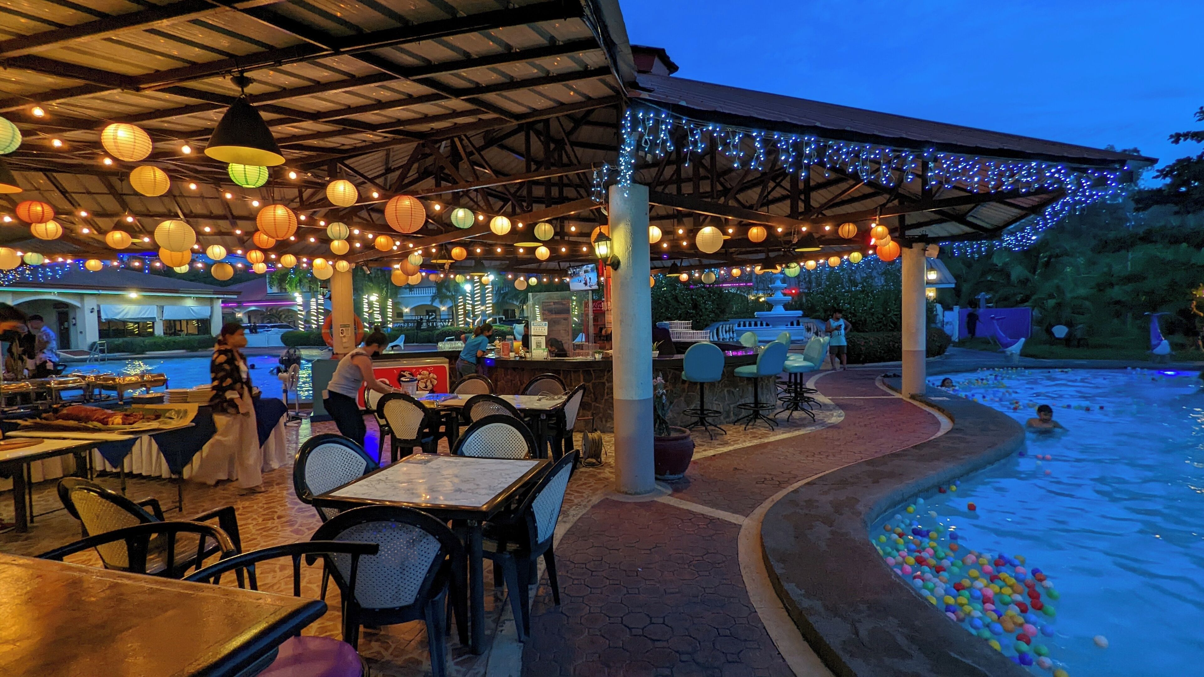 An outdoor dining area with tables and chairs, a pool with colorful balls, and a covered seating area with string lights.