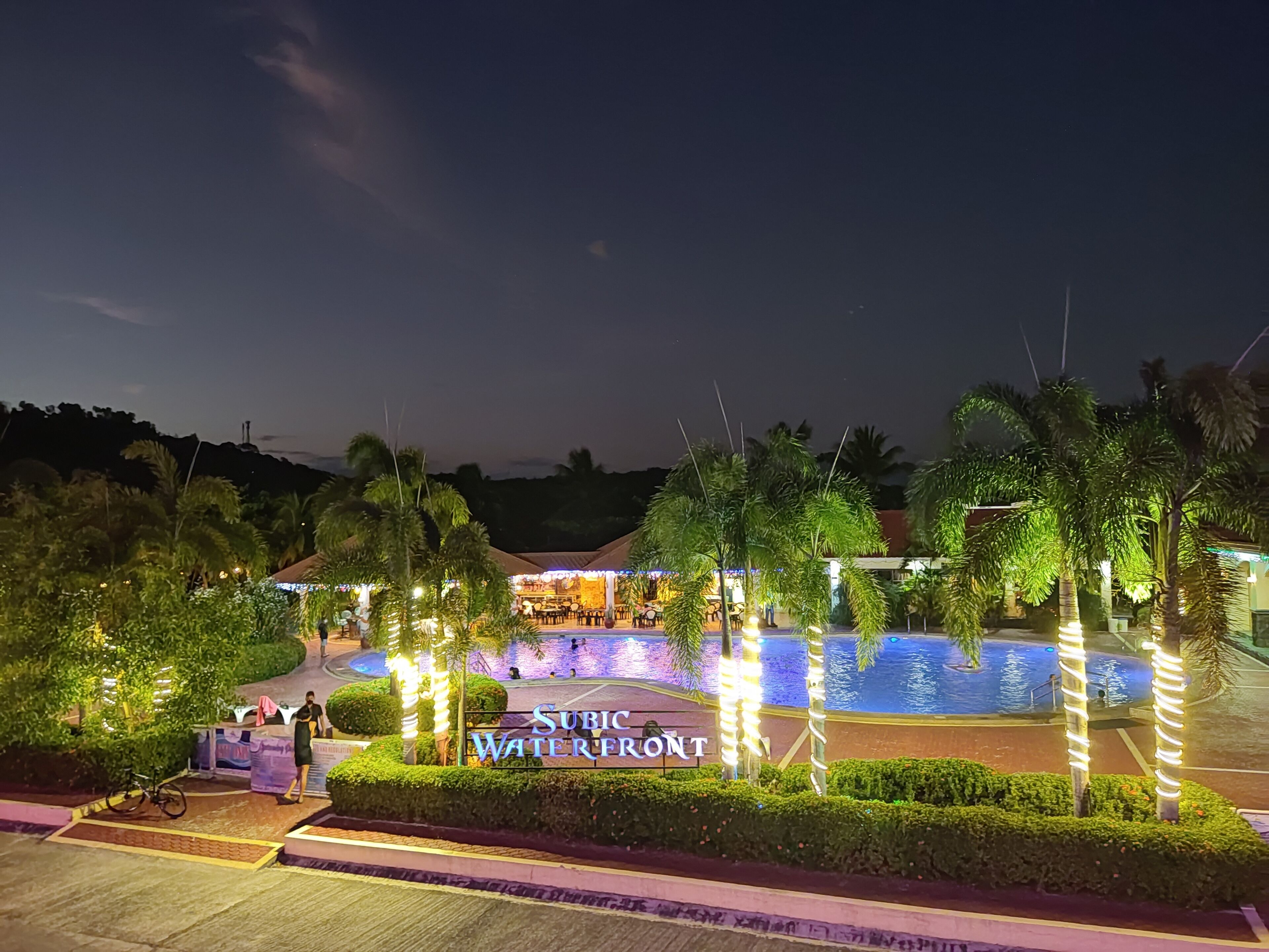 A nighttime view of a waterfront area with a pool, palm trees, and a sign reading "Subic Waterfront."