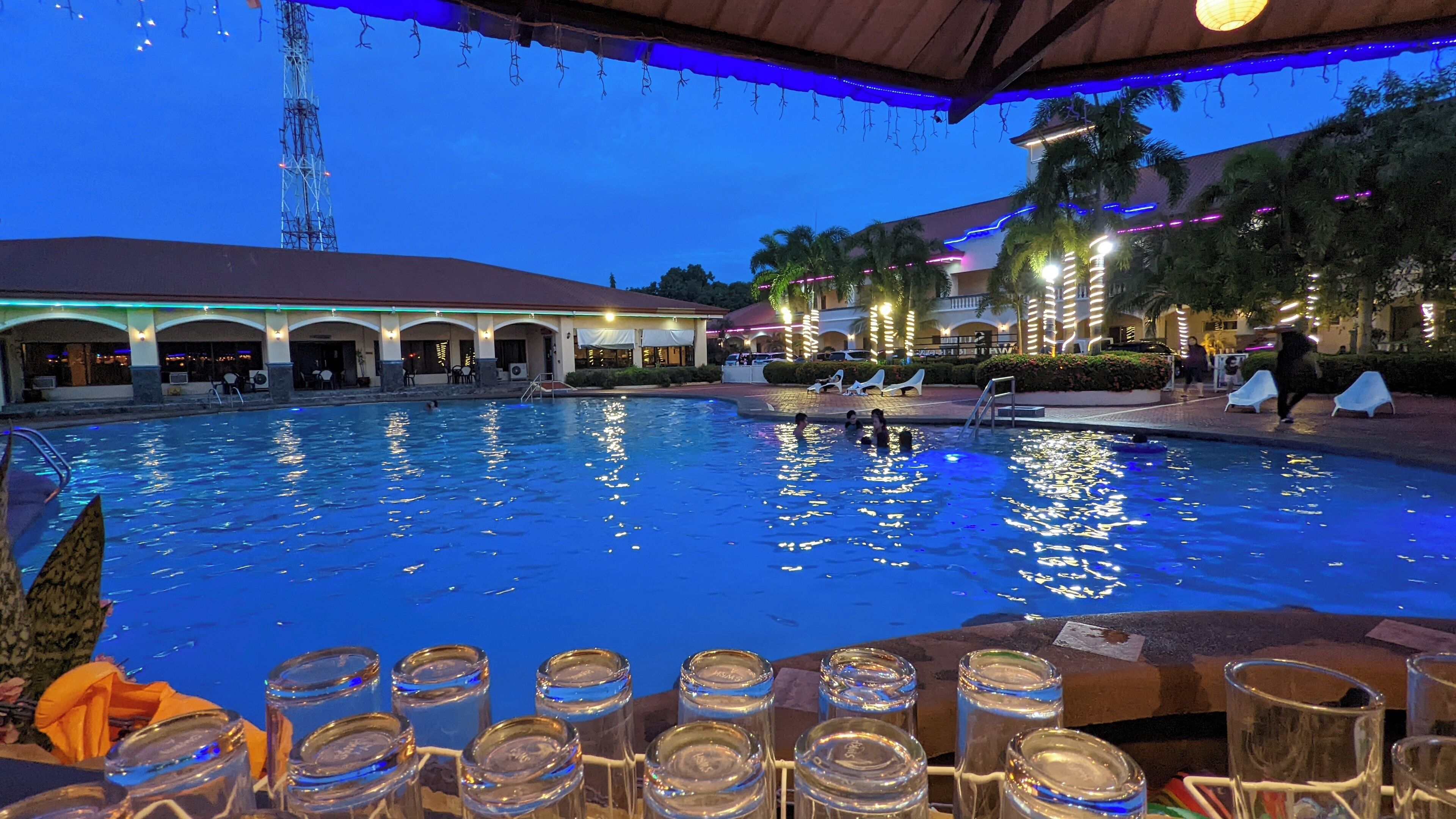 A swimming pool with lounge chairs and a covered area, surrounded by buildings with arches and palm trees.