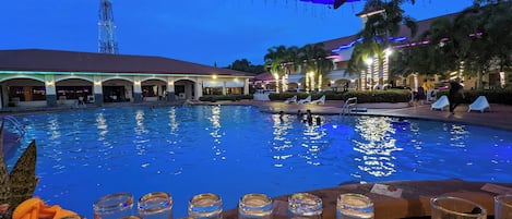 A swimming pool with lounge chairs and a covered area, surrounded by buildings with arches and palm trees.