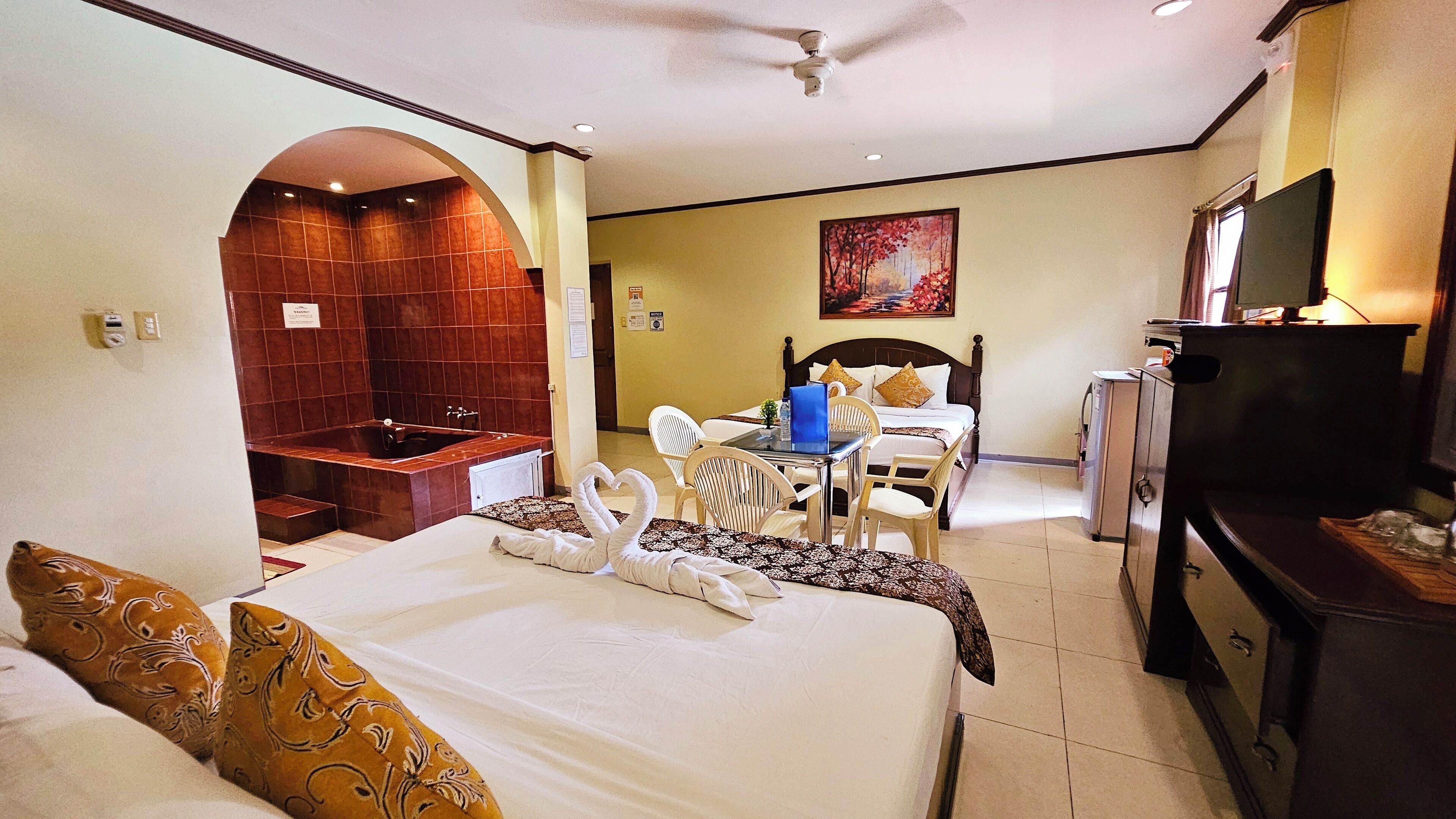 A hotel room with a large bed, a desk with chairs, a small table, a TV, a wardrobe, and a bathroom with a bathtub.