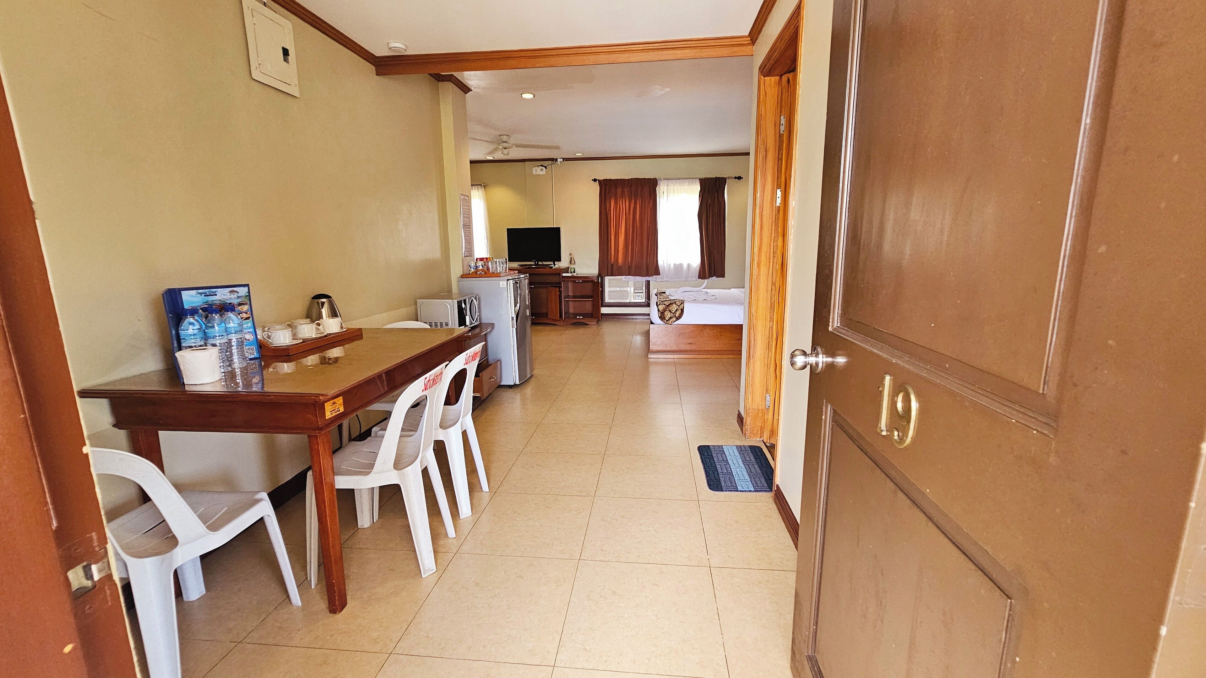 A hotel room with a wooden door, a bed, a desk with a computer, a refrigerator, and a microwave.