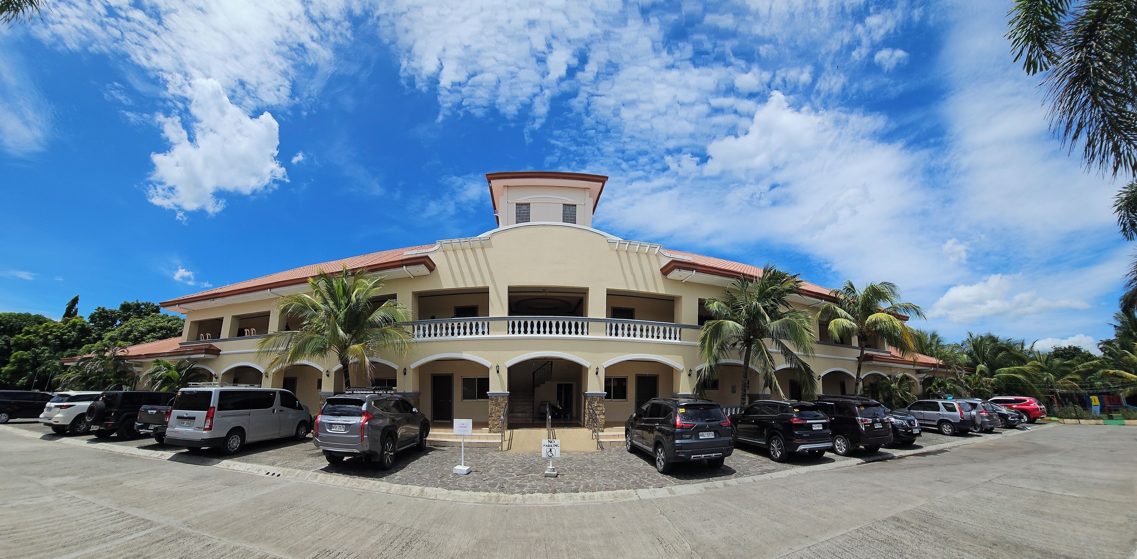 A multi-story building with a central balcony, parking lot, and surrounding palm trees.