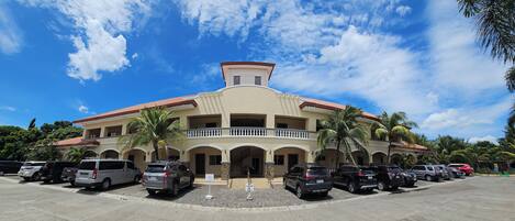 A multi-story building with a central balcony, parking lot, and surrounding palm trees.