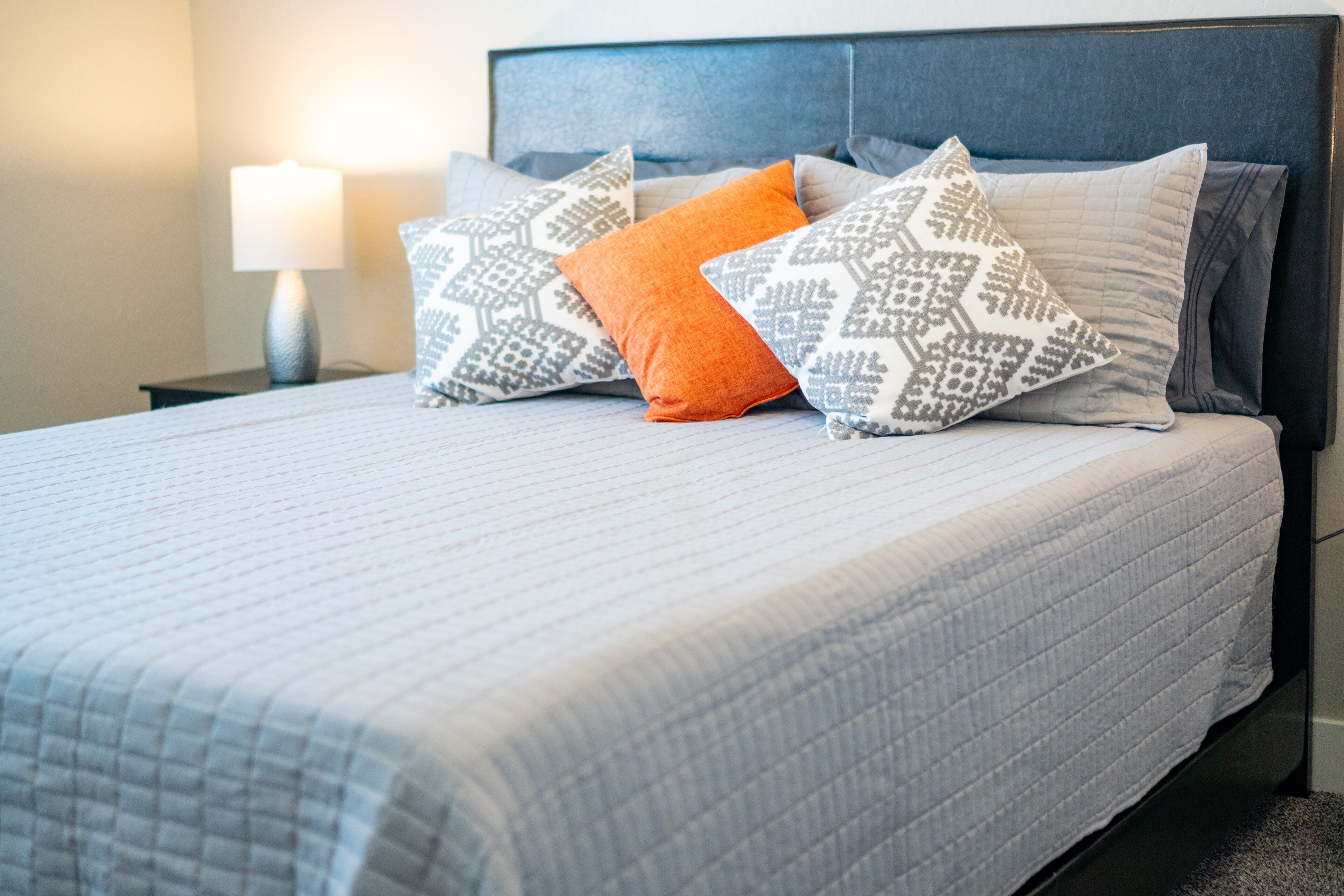Un lit bien fait, avec une couvre-lit à motifs et plusieurs oreillers, dont un oreiller orange bien visible.