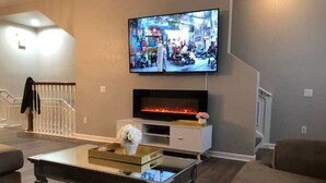 Ein modernes Wohnzimmer mit einem wandmontierten Flachbildfernseher, einem Kamin darunter und einem Glastisch in der Mitte.