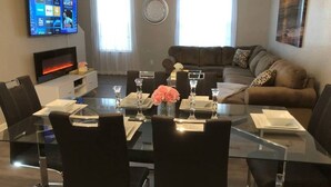 Ein modernes Wohnzimmer mit einem Glastisch für vier Personen, einem braunen Ledersofa, einem flachbildfernseher an der Wand und einem großen Fenster mit Vorhängen.