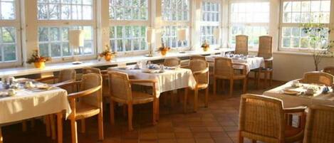 Un espace repas comprenant plusieurs tables et chaises, de grandes fenêtres et un sol carrelé.