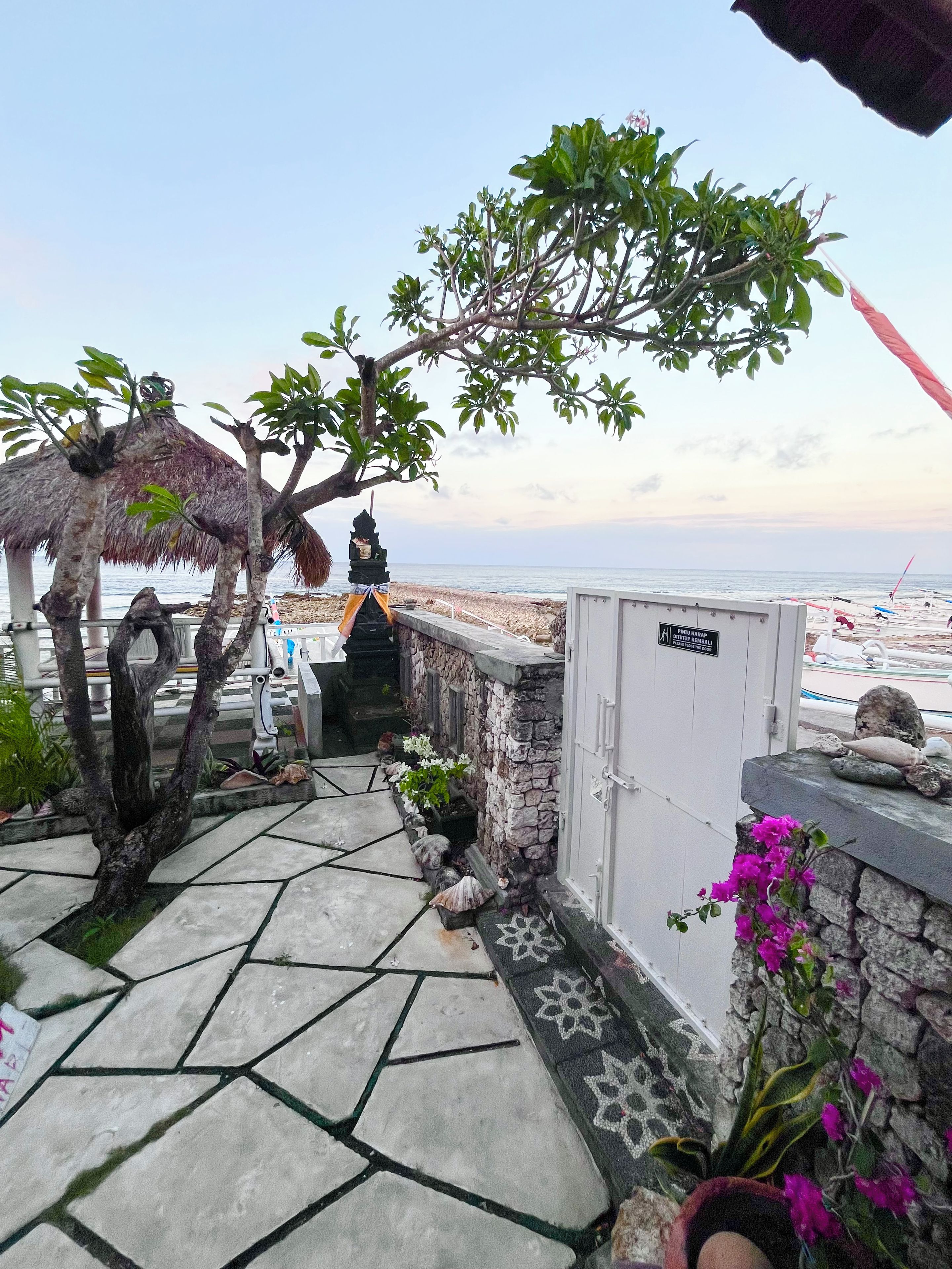 A stone pathway leading to a beach area with a thatched-roof structure, a small shrine, and a view of the sea.