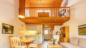 A spacious living area with a wooden balcony, a dining table with chairs, a sofa, and a kitchen in the background.