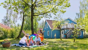 Une famille profite d’un pique-nique sur une couverture, dans un espace verdoyant, avec une maison bleue en arrière-plan.