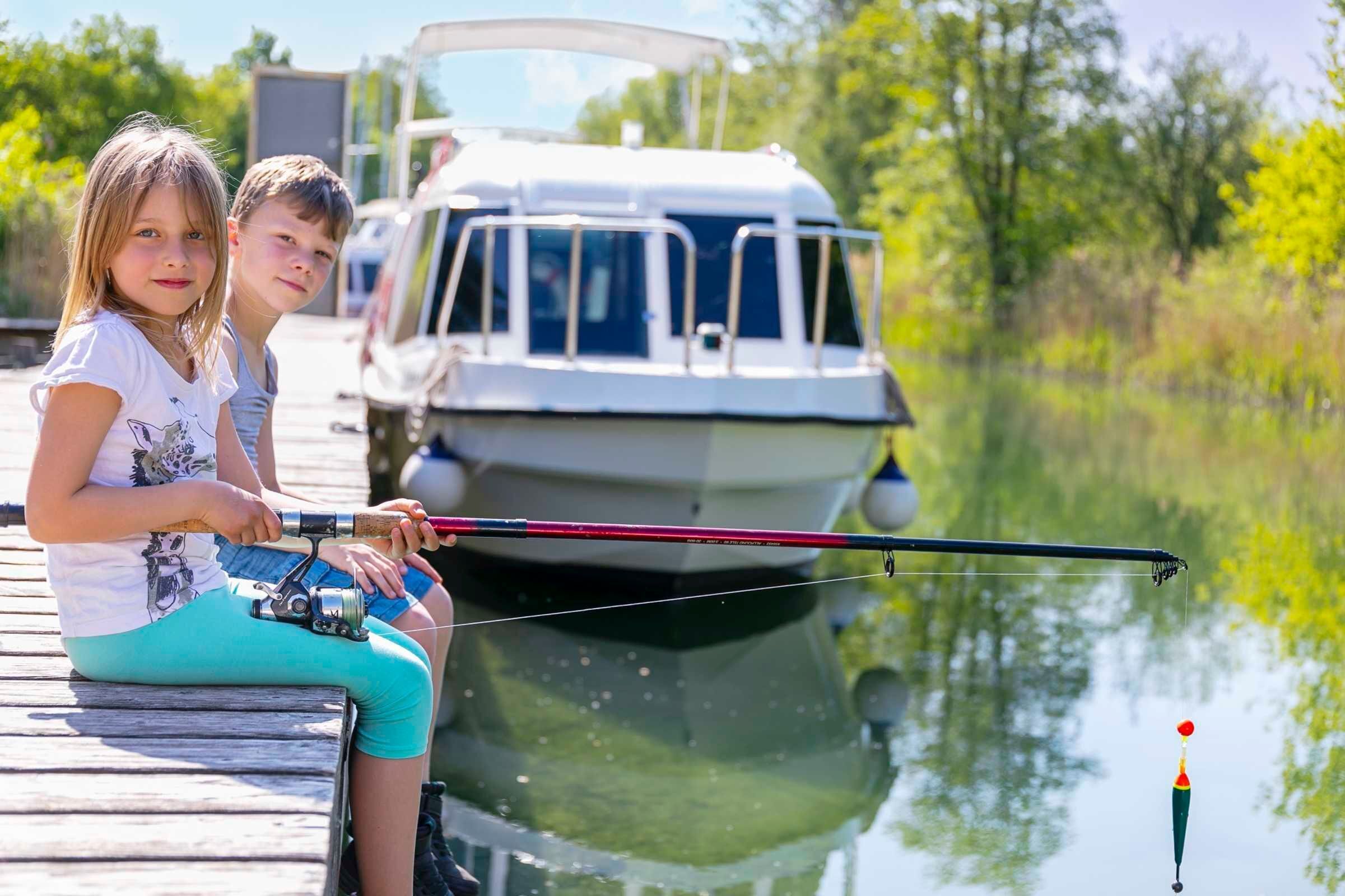 Zwei Personen angeln von einem Holzsteg, im Hintergrund ein Boot.