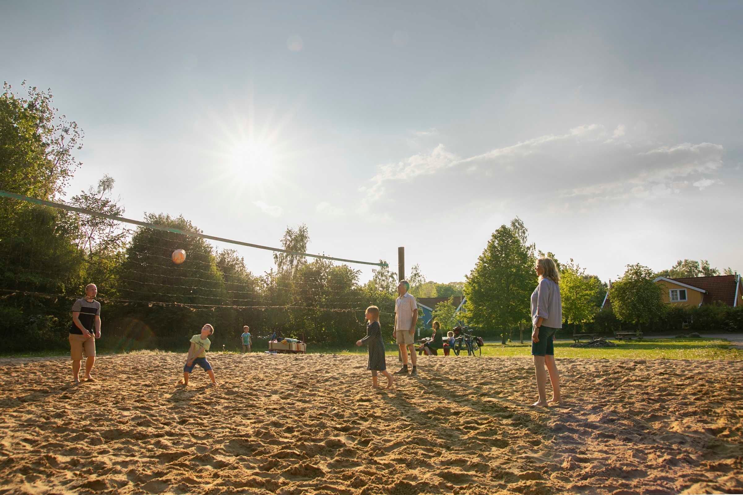 Un match de volley-ball se déroule sur un terrain sablonneux, délimité par un filet, et entouré d’arbres et de maisons.