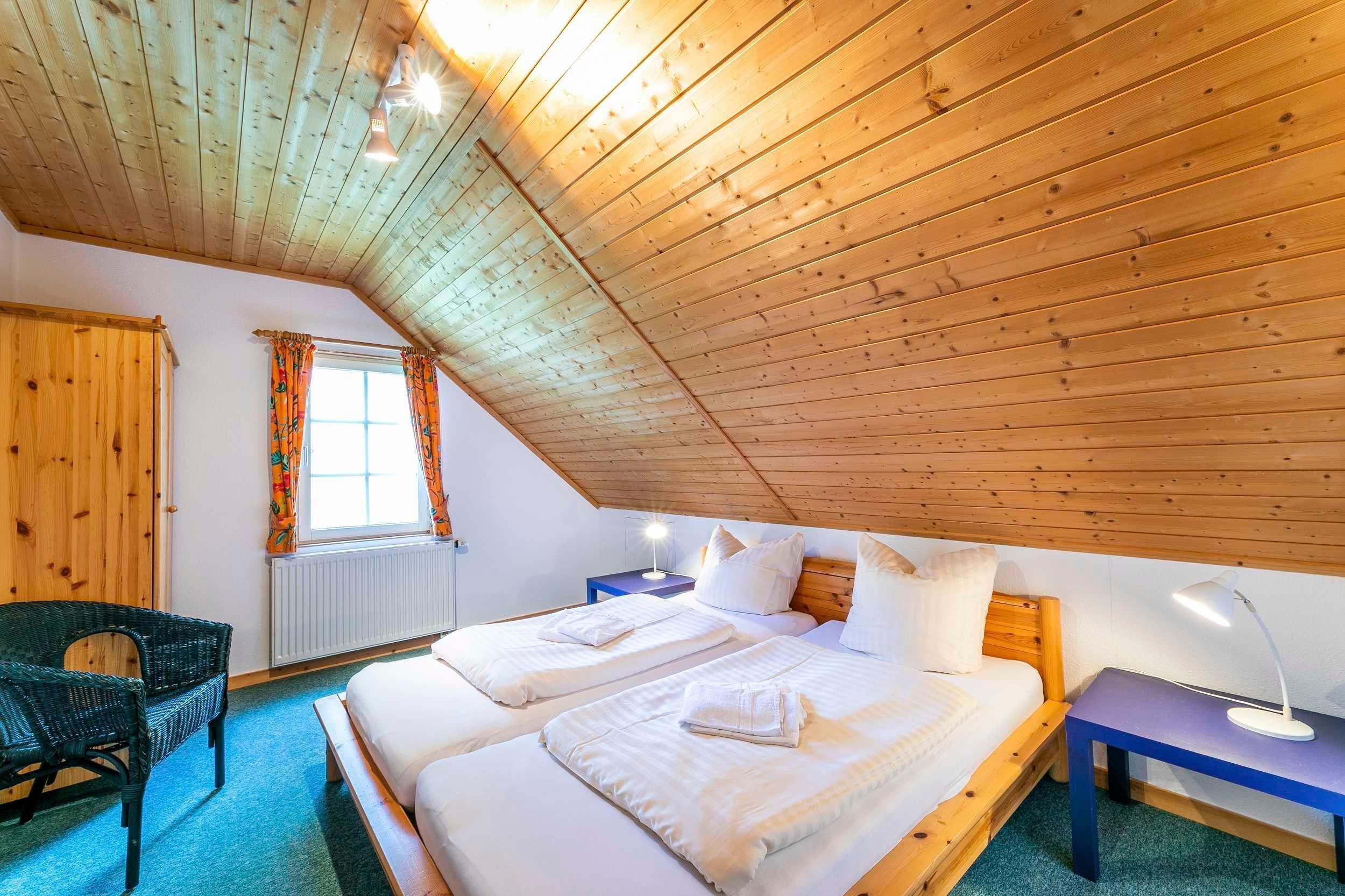 A cozy room with wooden ceiling beams, two beds with white linens, a wicker chair, and a small table with a lamp.