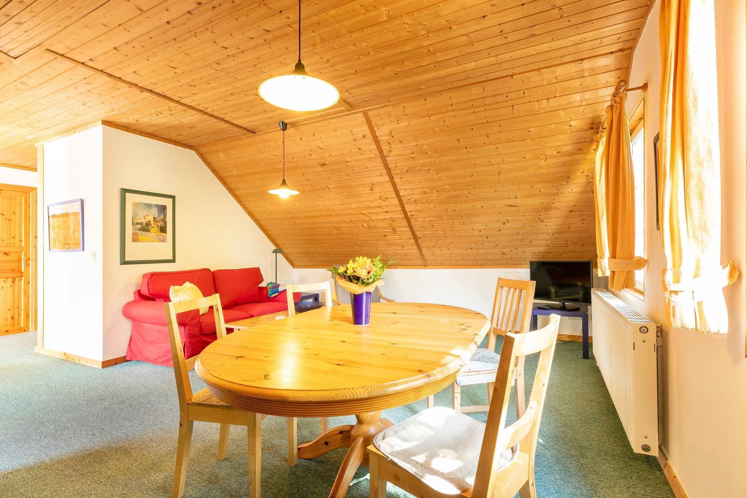 Ein gemütliches Zimmer mit Holzbalkendecke, einem runden Holztisch, einem roten Sofa und einem Fernseher.