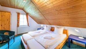 Une pièce chaleureuse avec des poutres apparentes en bois au plafond, deux lits avec du linge de lit blanc, un fauteuil en osier, une petite table avec une lampe et une fenêtre avec des rideaux.