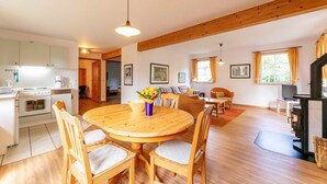 Ein gemütliches, helles Wohnzimmer mit Esstisch und Stühlen aus Holz, einem Kamin und einer Küchenzeile mit weißen Schränken und Geräten.
