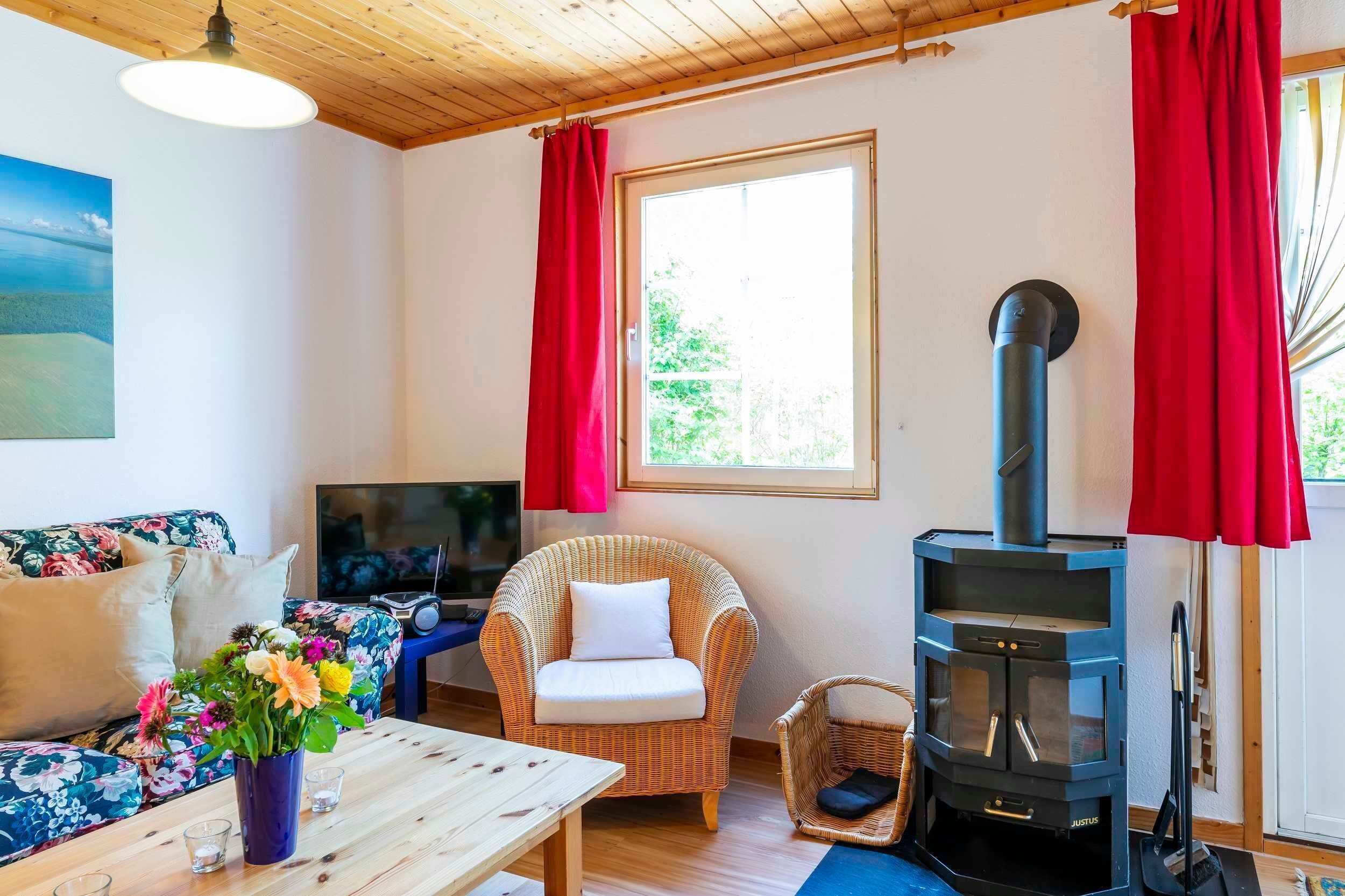 Una habitación acogedora con un sofá, una silla de mimbre, una mesa con un florero, una chimenea y una ventana con cortinas rojas.