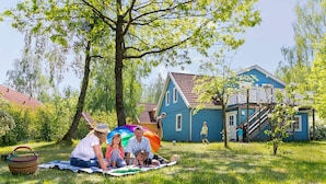 Une famille profite d’un pique-nique sur une couverture, dans un espace verdoyant, avec une maison bleue en arrière-plan.