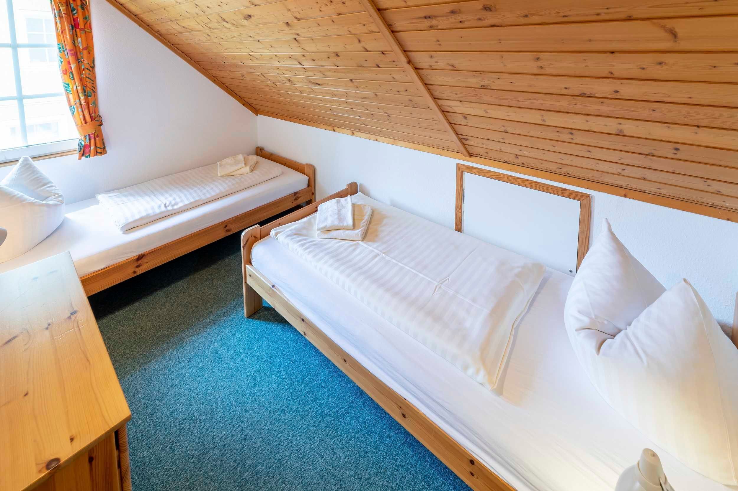 A small, cozy room with two single beds, a wooden desk, and a window with curtains.