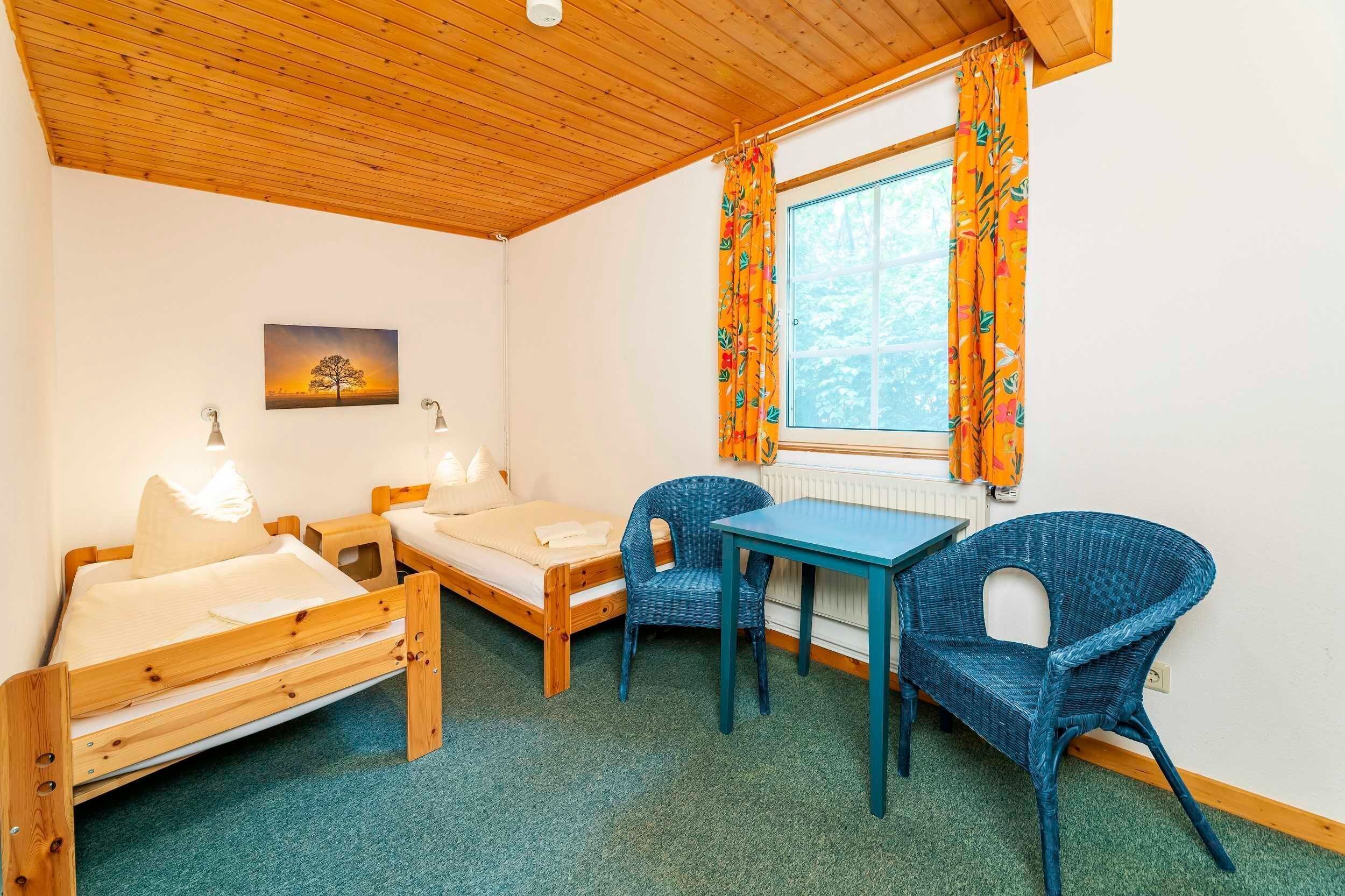 Een kleine kamer met twee houten bedden, een tafel, twee rieten stoelen, een raam met gordijnen, en een schilderij aan de muur.