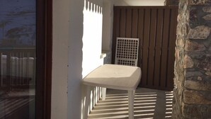 Un pequeño balcón con una mesa y una silla blancos, una ventana con vistas y un muro de piedra.