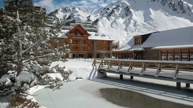 Un complejo turístico en la montaña cubierto de nieve, con edificios de madera y un puente sobre un pequeño cuerpo de agua.