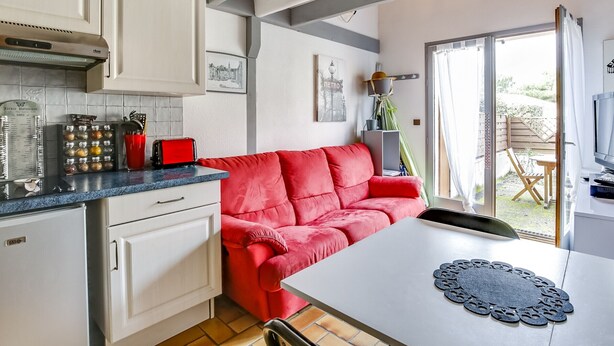 Eine kleine Küche mit weißen Schränken, einem roten Sofa und einem Esstisch mit Platzdeckchen.