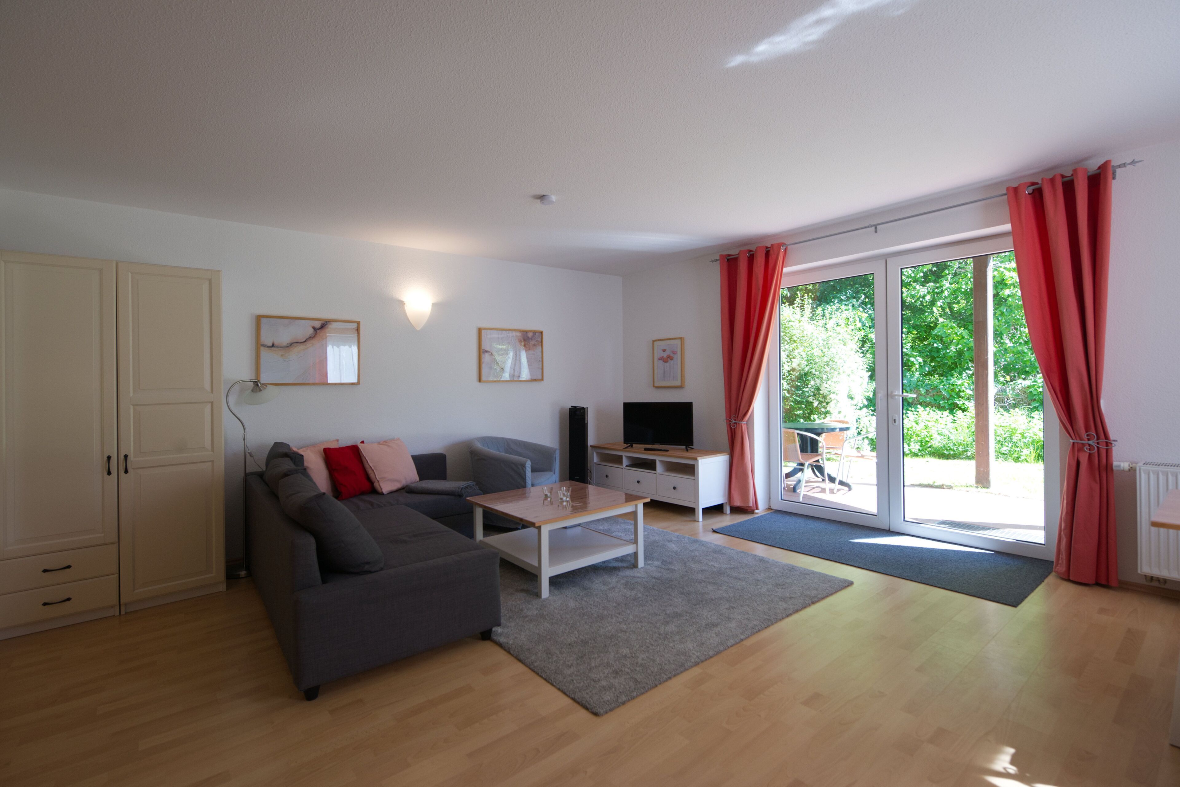 A modern living room with a sofa, coffee table, and TV, a sliding glass door to a garden, and a white cabinet.