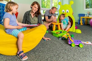 A family in a playroom with a child on a bean bag, another child playing with toys, and an adult supervising. - 2-room vacation apartment, 2 (+2) persons, ground floor (13) - Müritzparadies - Feriendorf (Rechlin)