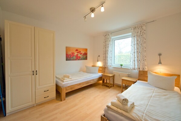 A bedroom with two single beds, a wooden wardrobe, a small bedside table, a window with curtains, and a painting on the wall.