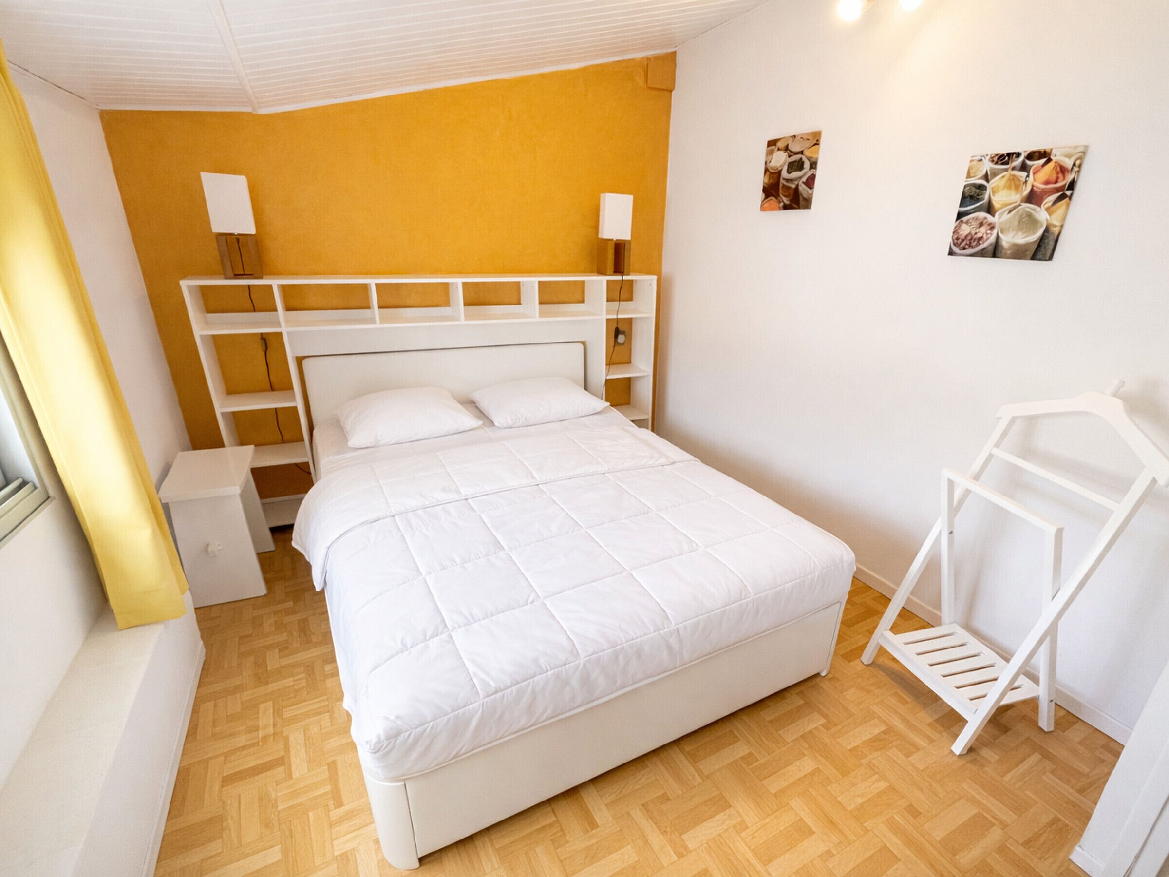Een wit bed in een kamer met een houten vloer, een geel accentmuur en een witte wandplank.