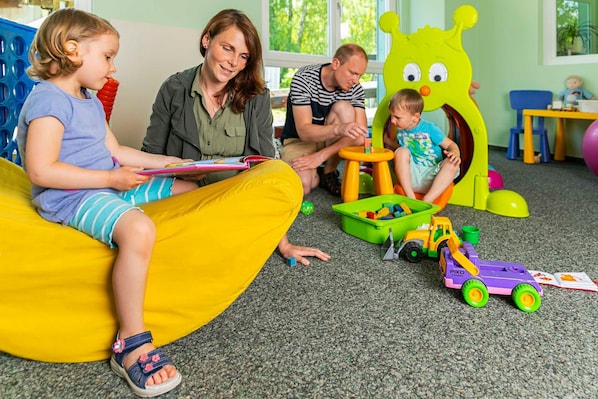 Eine Familie im Spielzimmer: ein Kind sitzt auf einem Sitzsack, ein anderes Kind spielt mit Spielzeug, und ein Erwachsener passt auf.