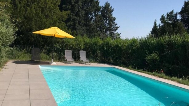 A rectangular swimming pool with clear blue water, surrounded by a tiled deck. There are two lounge chairs and a yellow umbrella providing shade.