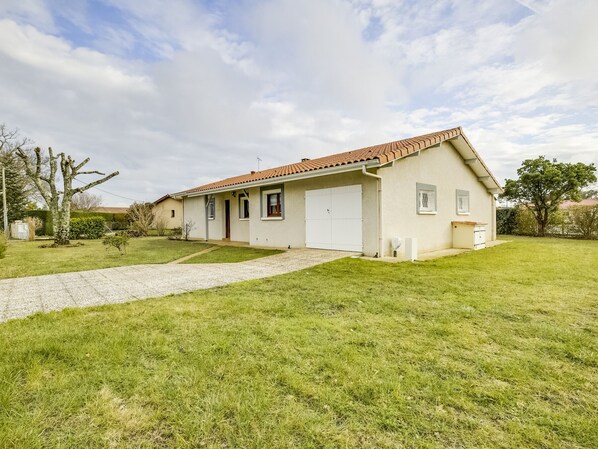 A single-story house with a tiled roof, a front door, and a garage. The property has a well-maintained lawn and a gravel driveway. - Cozy 3-Bedroom Home with Garden & BBQ, Near Beach and Lake, Biscarrosse (Biscarrosse)