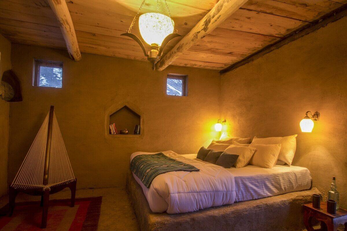 A cozy bedroom with a wooden ceiling, a bed with pillows, a small table with a bottle, and two wall-mounted lights.