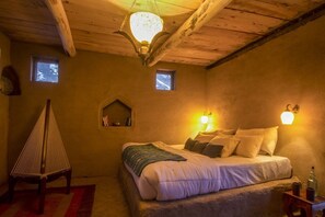 Un dormitorio acogedor con techo de madera, una cama con almohadas, una mesita con una botella y dos luces fijadas a la pared.