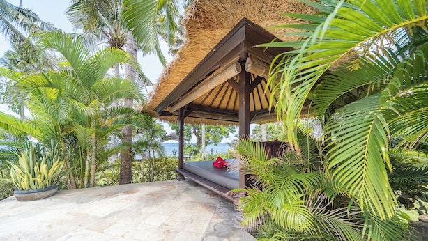 Un'area salotto all'aperto riparata da un tetto di paglia, circondata da rigogliosa vegetazione tropicale e palme, con vista su un'ampia distesa d'acqua.