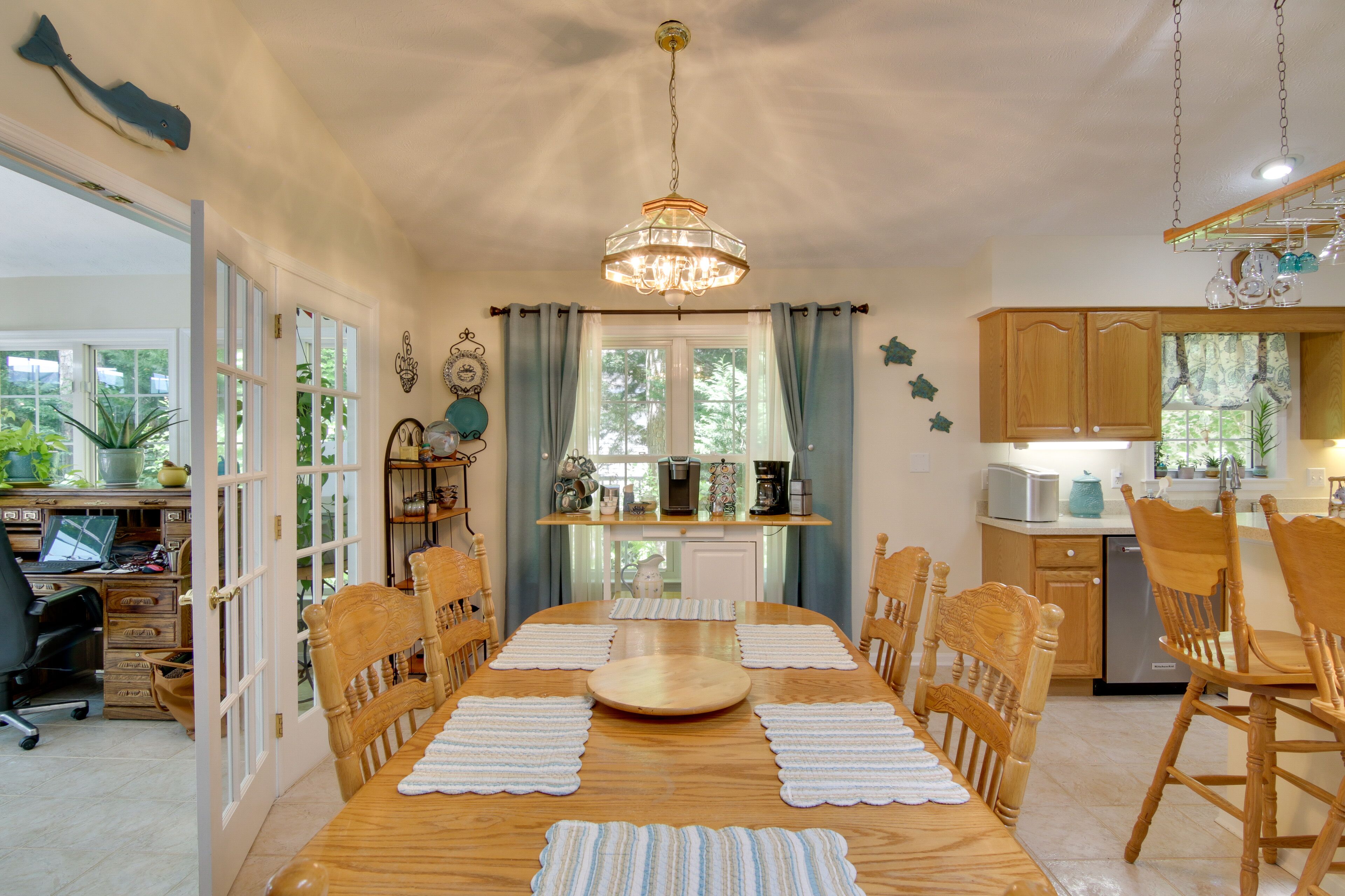 A dining room with a wooden table, chairs, and a chandelier. There is a kitchen area with cabinets and appliances visible through an open door.