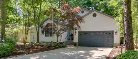 Une maison de plain-pied avec un garage, entourée d’arbres adultes et d’un jardin aménagé.