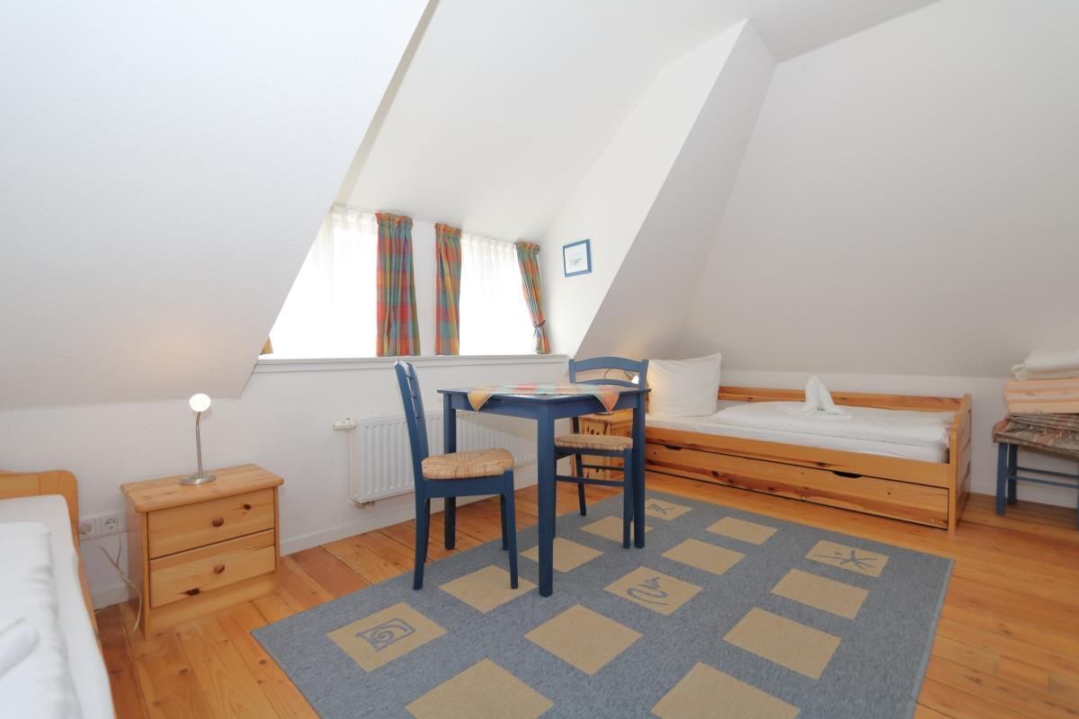 A small, well-lit room with a sloped ceiling, a single bed, a desk with a chair, and a window with curtains.