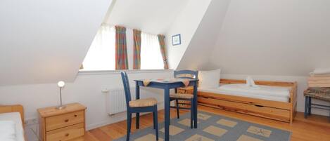 A small, well-lit room with a sloped ceiling, a single bed, a desk with a chair, and a window with curtains.