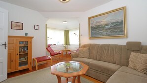 A living room with a beige sofa, a glass-top coffee table, a wooden cabinet, a red bench, and a painting of a seascape. - Haus Sonnenschein, Germany (Sylt)