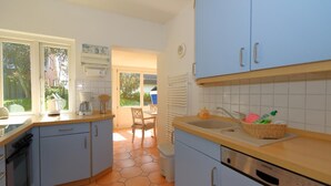 A kitchen with light blue cabinets, a dishwasher, and a window overlooking a garden. - Haus Sonnenschein, Germany (Sylt)