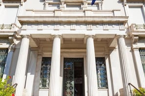 A classical building with a central entrance flanked by columns, large windows with metal grilles, and a decorative pediment above.