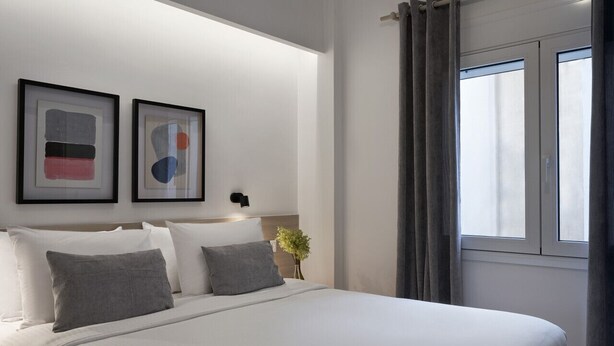 A modern hotel room with a large bed, two framed abstract artworks, a bedside table with a vase of flowers, and a window with curtains. - Parthenon 2BD Apartment (Athens)