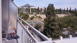 A balcony with a view of ancient ruins, a hilltop, and a modern building in the background. - Parthenon 2BD Apartment (Athens)