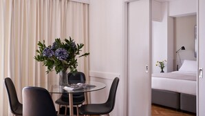 A modern hotel room with a glass dining table, two black chairs, a vase of flowers, a bed with a white mattress and pillows, and a small bedside table with a lamp. - Parthenon 2BD Apartment (Athens)