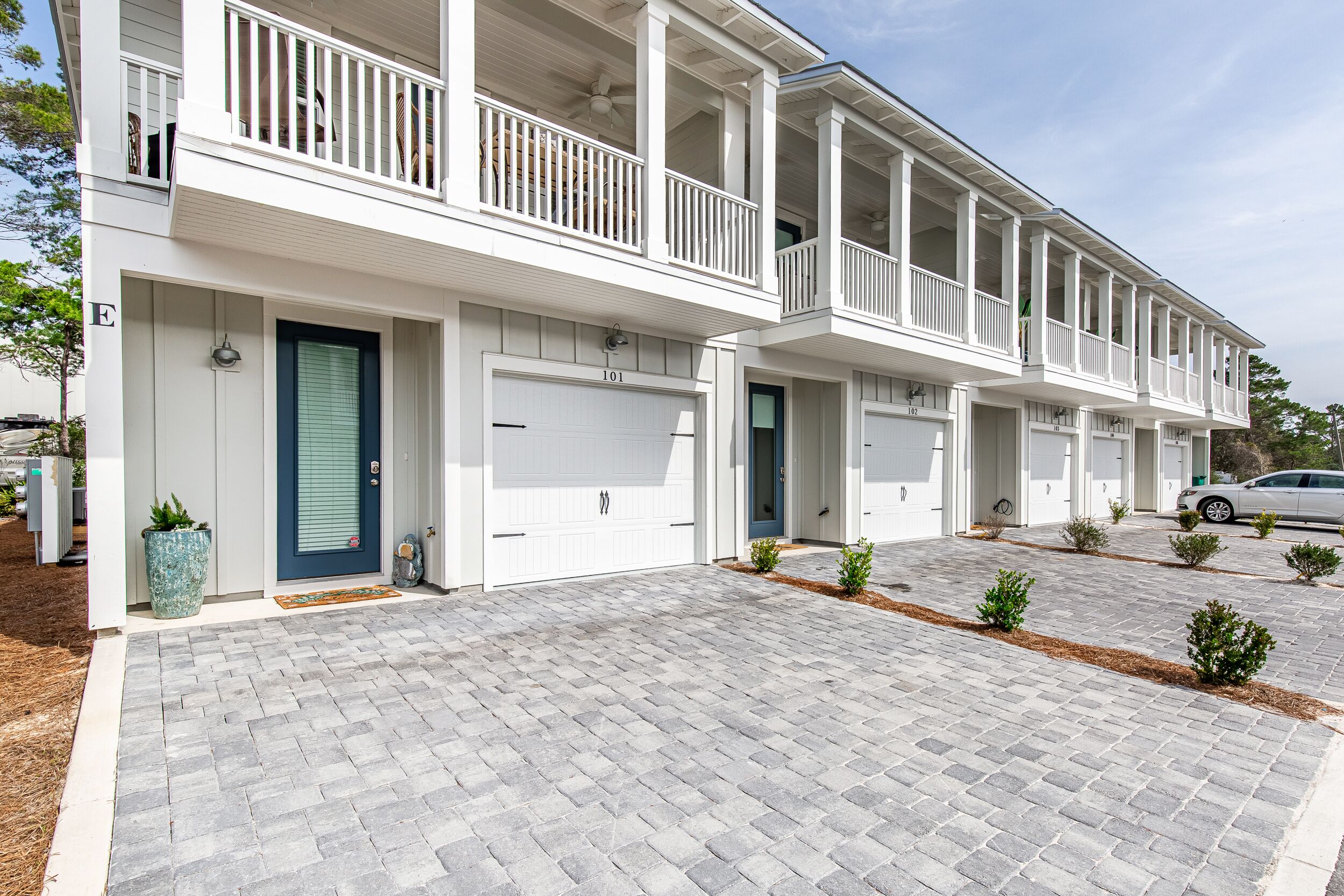 Une rangée de maisons de ville blanches à deux étages, avec des garages attenants, une allée pavée en briques et de petits arbustes dans le jardin.